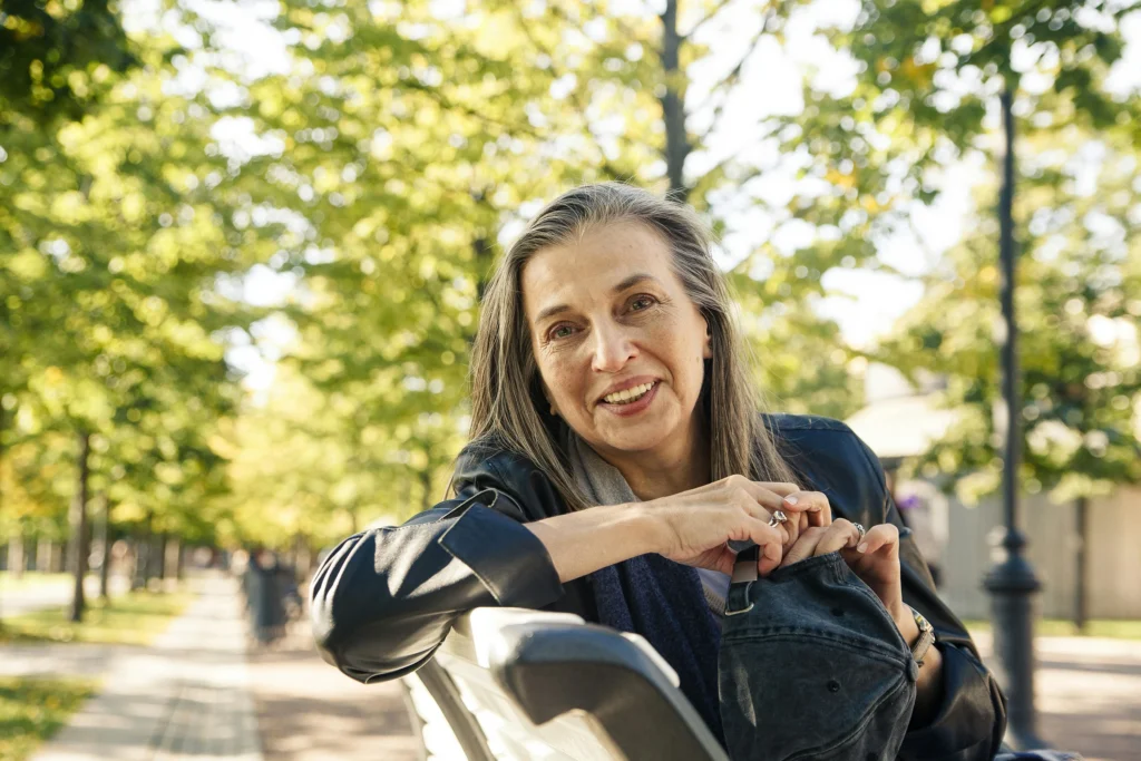 Woman smiling while sitting on a park bench on a sunny, tree-lined path.