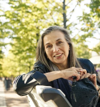 Woman smiling while sitting on a park bench on a sunny, tree-lined path.